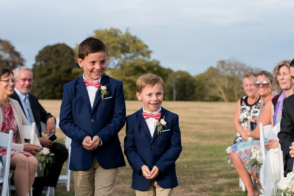 Two boys in navy suits walk down an aisle outdoors at a wedding ceremony