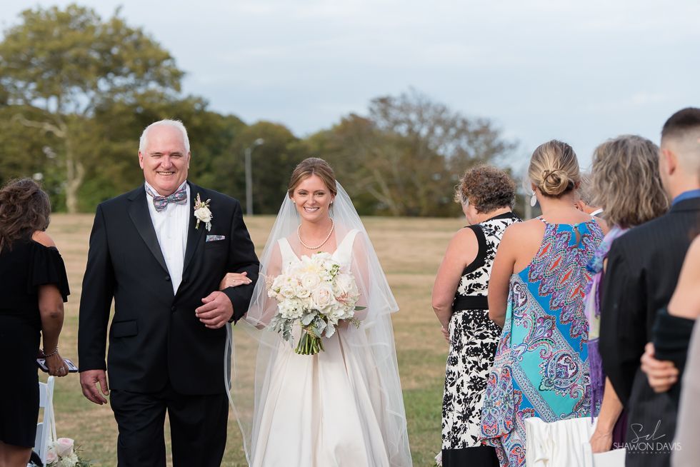 Bride in white gown walks with an older man in a tuxedo at an outdoor wedding ceremony