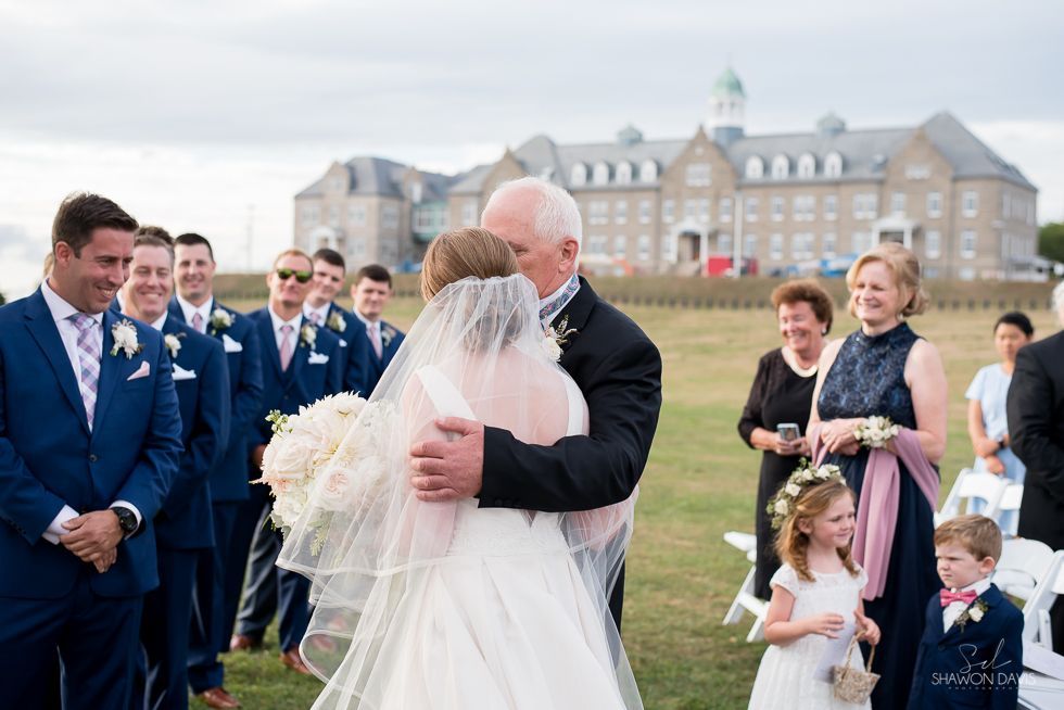 Wedding couple embracing outdoors with guests gathered near a large stone building