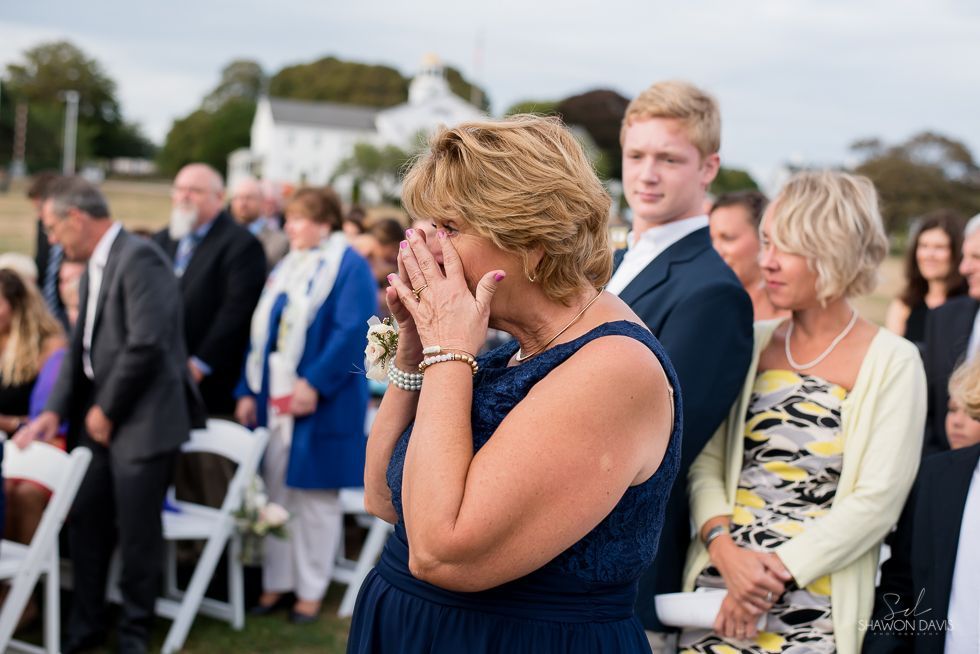 Woman in navy dress wipes tears at outdoor gathering, with seated guests in the background