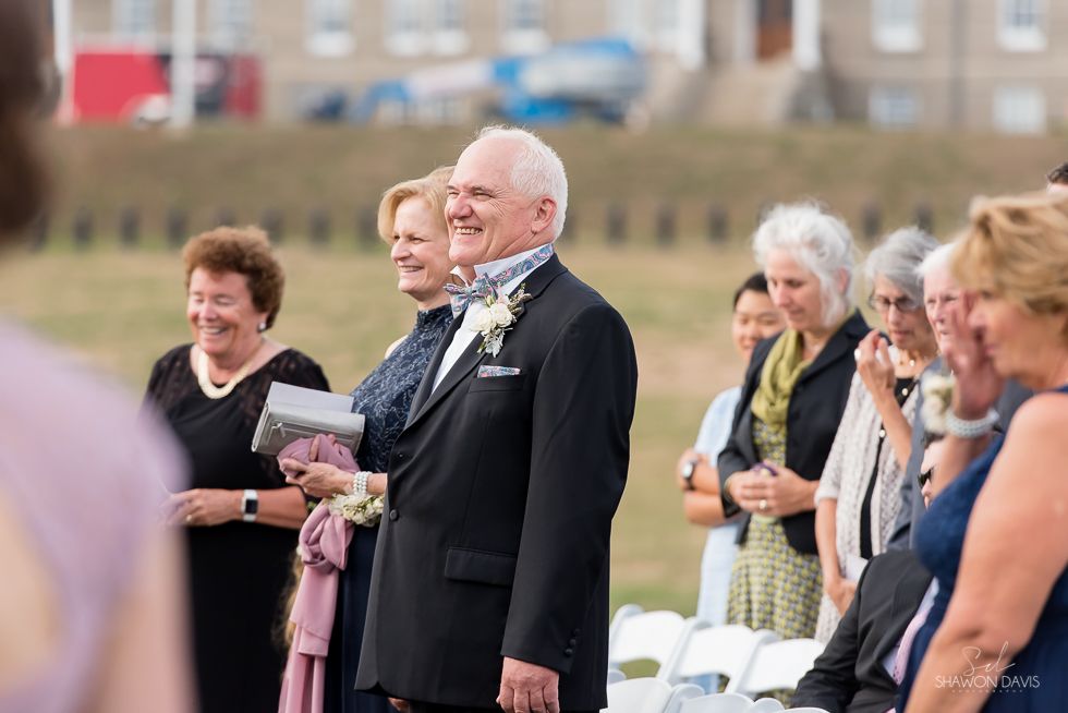 People standing outdoors at a wedding ceremony, with a smiling man in a black suit holding pink flowers.