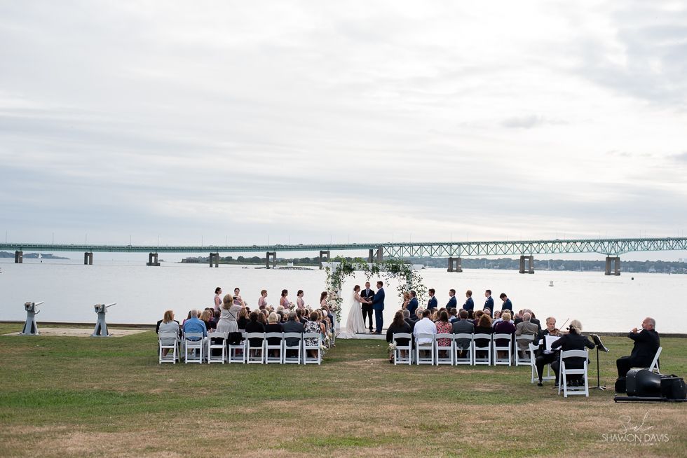 People seated outdoors by the water, with a long bridge in the background.