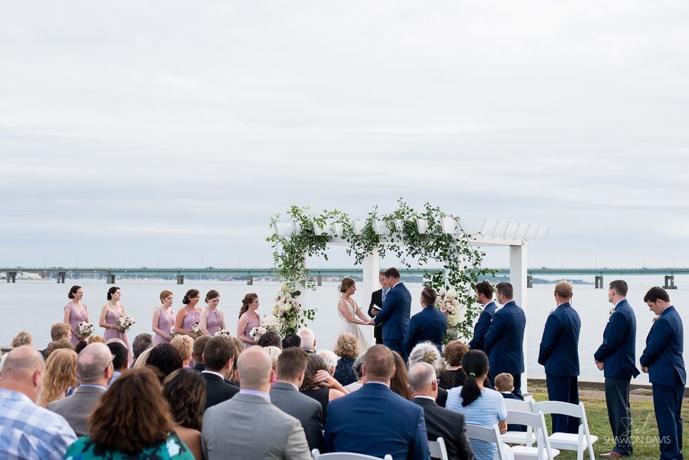 Wedding ceremony by the water with guests seated and a floral arch under a cloudy sky