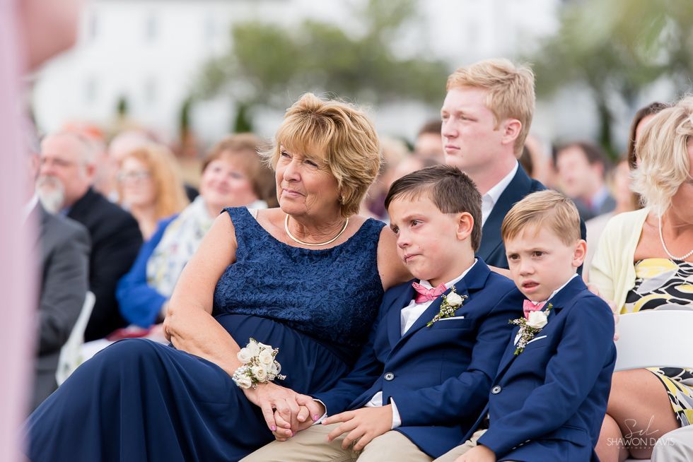 Two boys in blue suits sit beside a woman in a navy dress at an outdoor ceremony