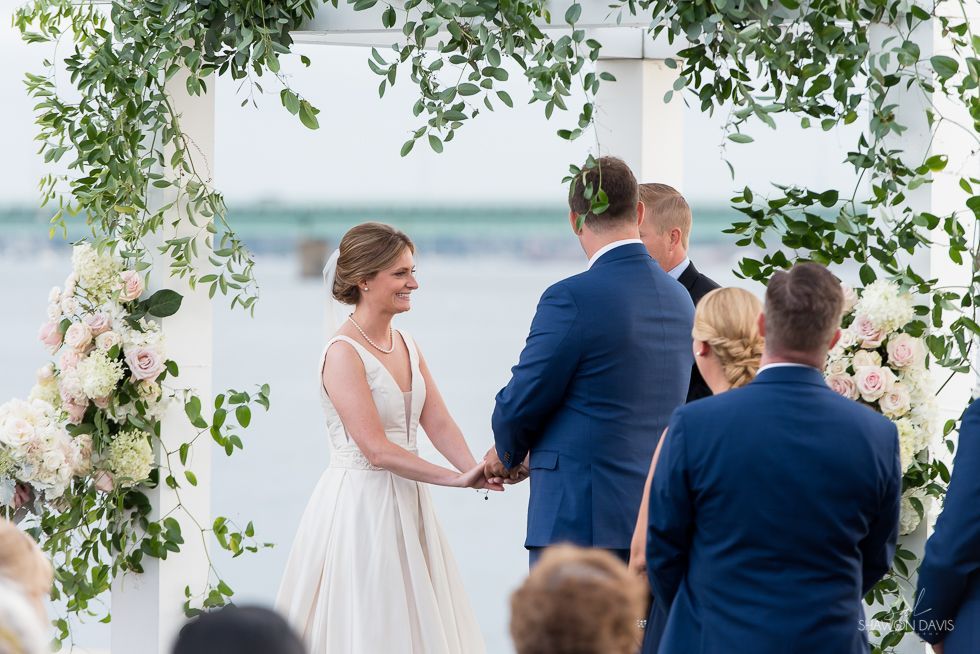 Bride and groom hold hands under a floral arch at an outdoor waterfront wedding ceremony