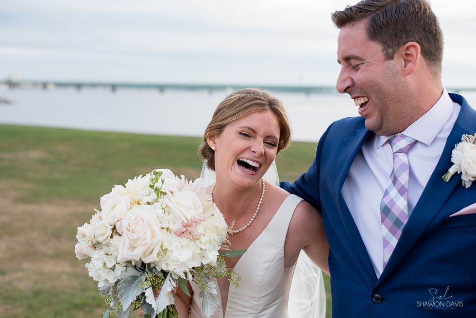Newlyweds laughing outdoors by the water, bride holding a white bouquet, groom in a blue suit