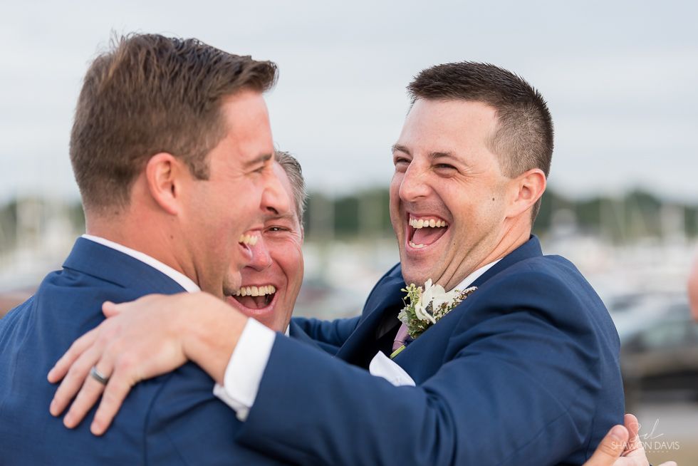 Three men in blue suits embrace and laugh outdoors at a waterfront celebration