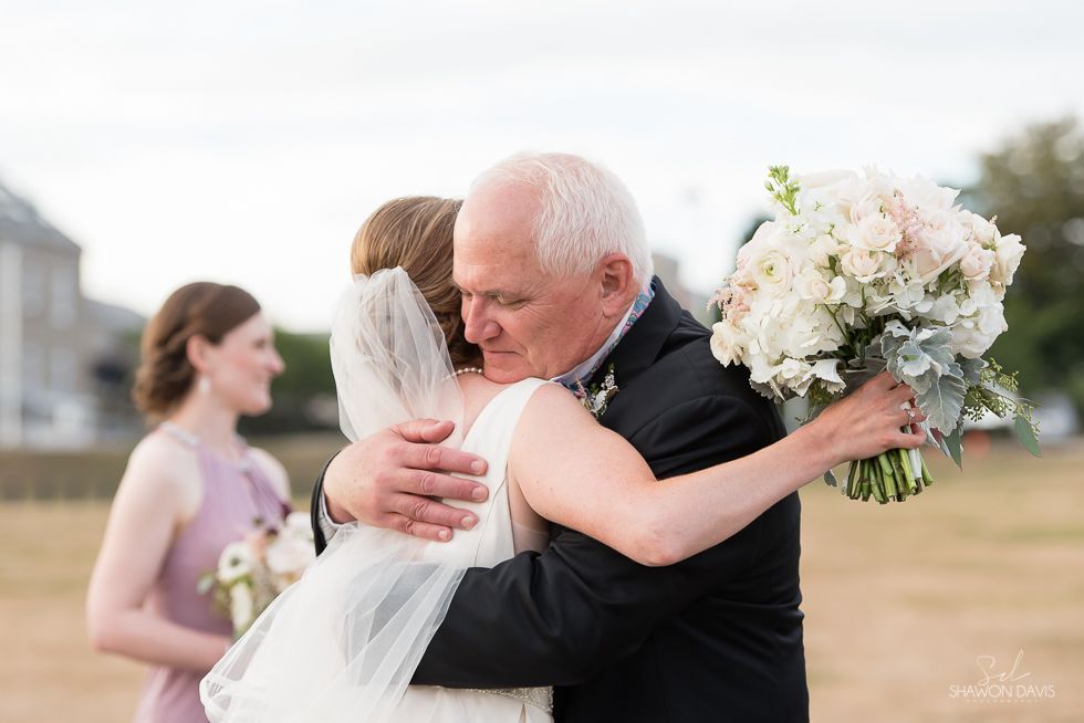 Woman in a white dress hugs an elderly man at an outdoor wedding while holding a bouquet.