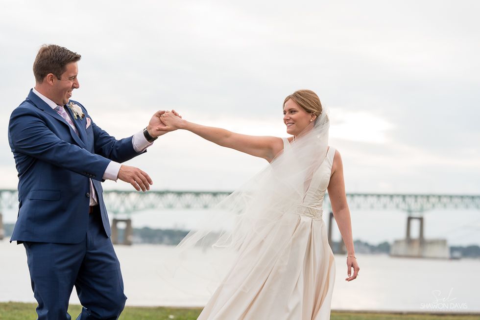 Bride and groom holding hands and dancing by a river with a bridge in the background