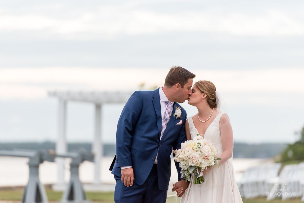 Bride and groom kiss outdoors by the water, with a white arbor in the background