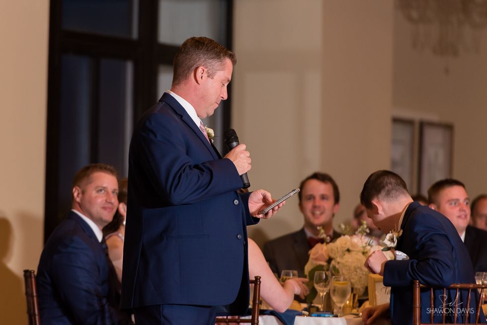 Man speaking into a microphone at a formal dinner, with seated guests and tables in the background.