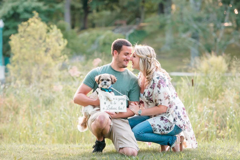 Couple kneeling in a grassy field, holding a small dog with a sign, embracing outdoors.