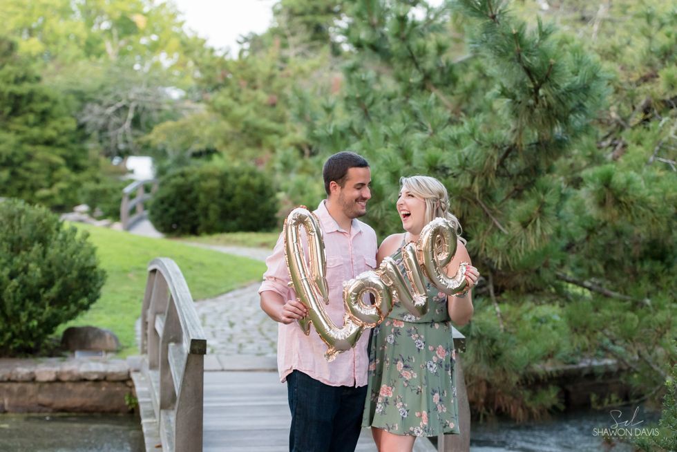 Couple holding rose-gold balloons on a garden bridge, smiling at each other.