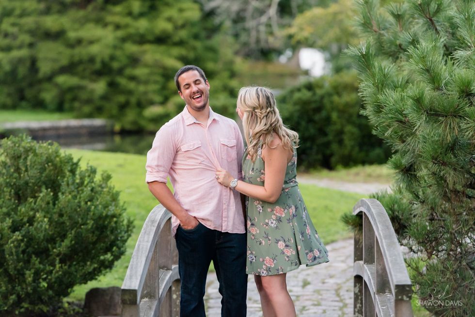Couple standing on a small bridge in a green park, smiling and embracing.