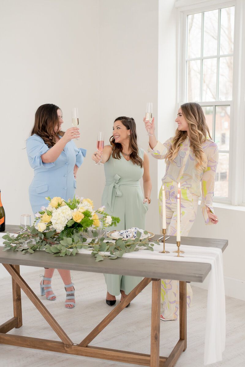 Three women toast with drinks beside a floral table in a bright room with large windows.