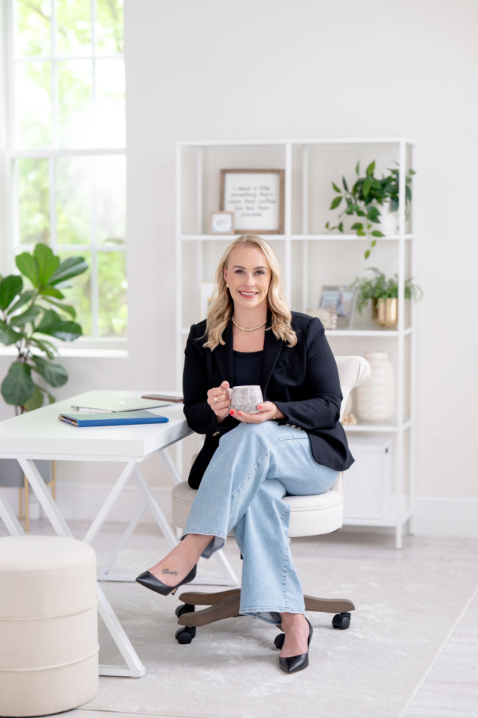 Woman in a blazer and jeans smiling at a desk in a bright home office with shelves and decor