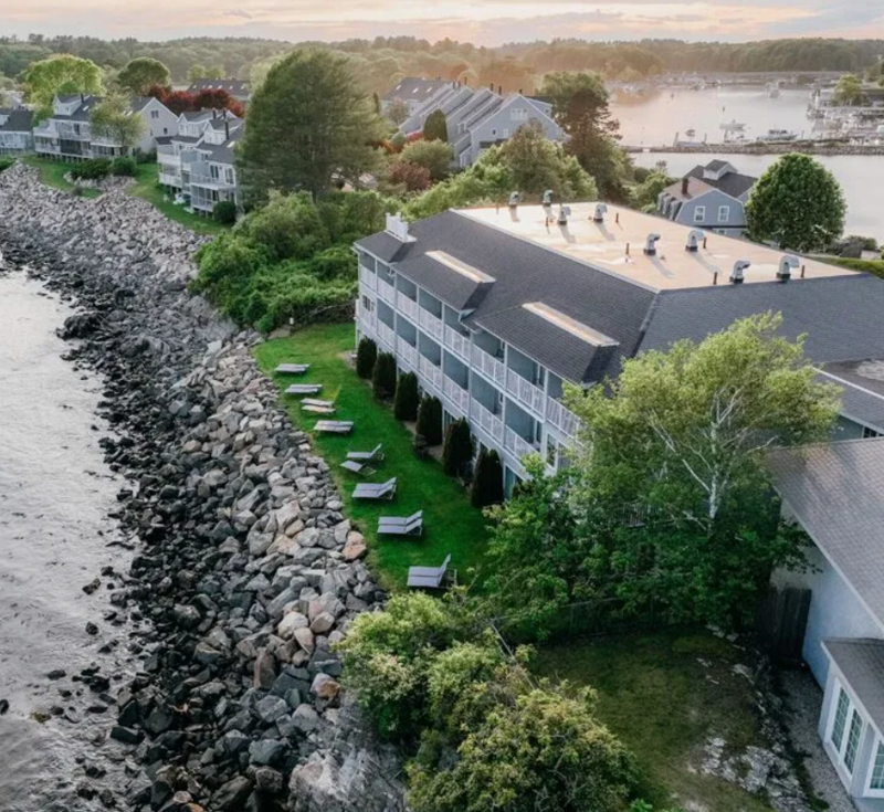 Aerial view of a waterfront building with lounge chairs beside a rocky shoreline and trees at sunset