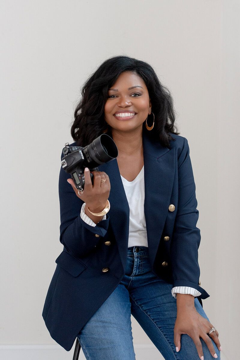 Woman seated against a light background, smiling and holding a camera while wearing a navy blazer and jeans.