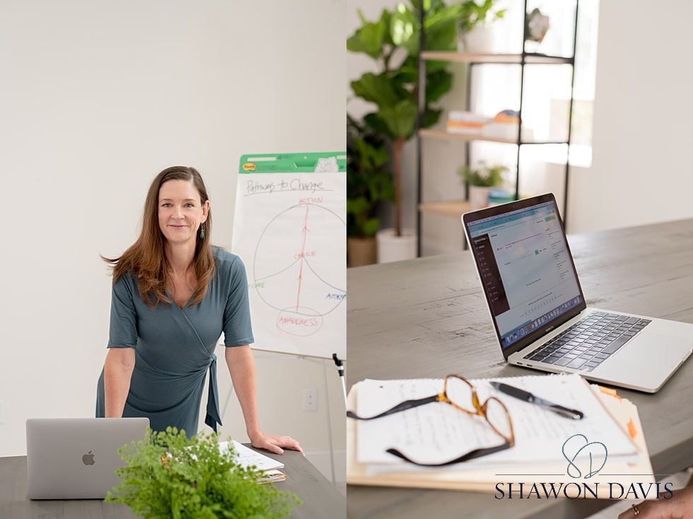 Woman standing by desk in bright office, with laptop, plant, monitor, and whiteboard behind her.