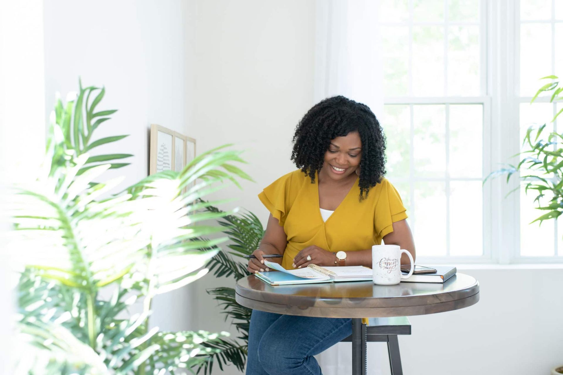 Woman in a yellow shirt writing at a table by a bright window with plants