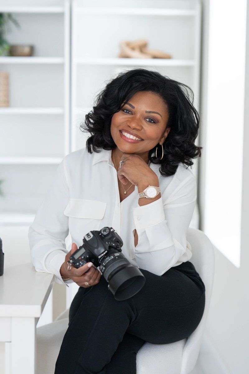 Woman in white blouse smiling while holding a camera in a bright white room