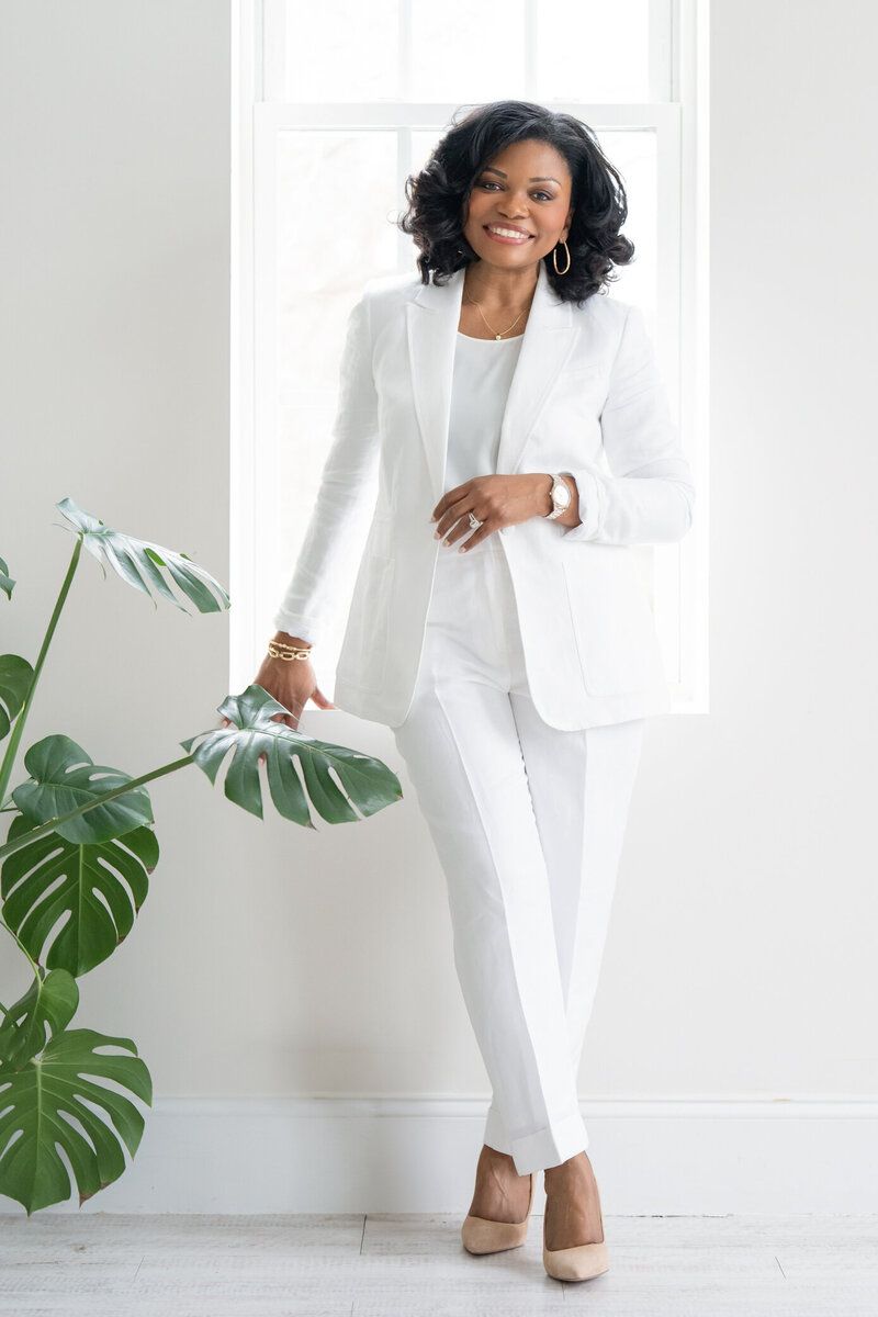 Woman in a white suit standing beside a leafy plant in a bright room
