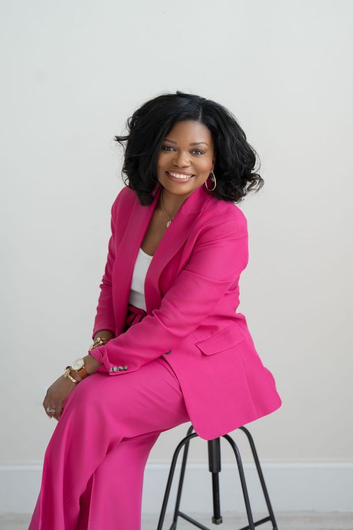 Smiling woman seated on a white couch, wearing a navy blouse and jeans against a light gray wall.