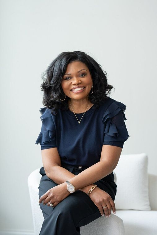 Smiling woman seated on a white couch, wearing a navy blouse and jeans against a light gray wall.