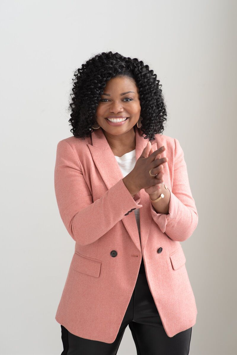 Smiling person in a pink blazer clapping against a plain light background