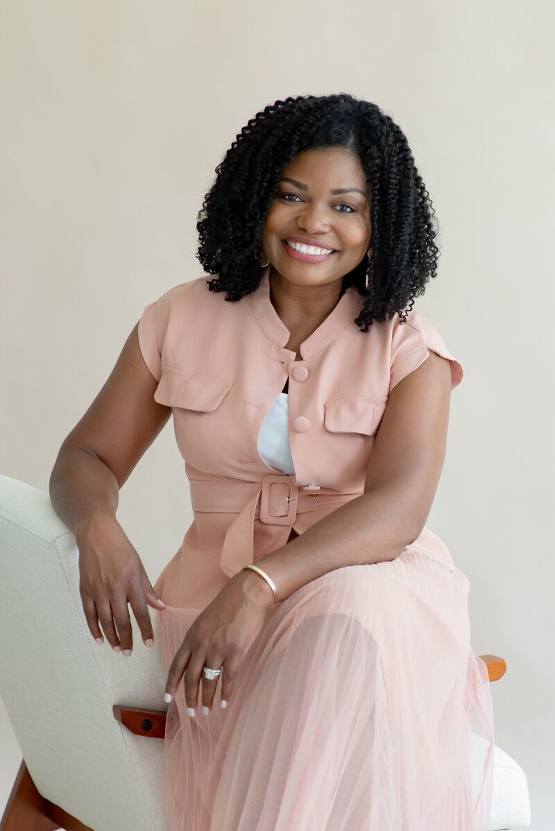 Smiling woman seated against a neutral wall, wearing a blush pink outfit and white top.