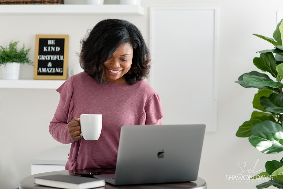 Person smiling at a laptop while holding a white mug in a bright home office