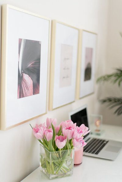 Framed art above a desk with a laptop and vase of pink tulips in a bright room