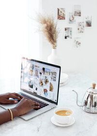 Hands typing on a laptop at a bright desk with coffee and a glass kettle nearby