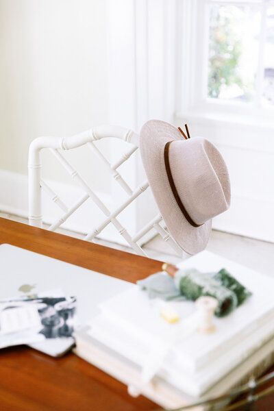 Beige hat hanging on a white chair beside a sunlit table with books and flowers