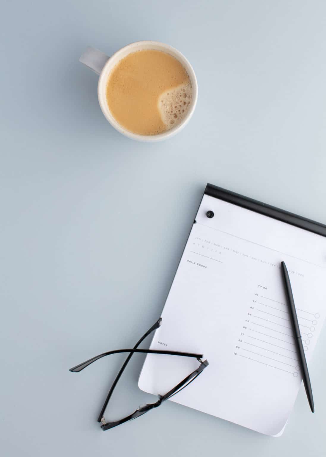 Coffee cup, eyeglasses, pen, and a notepad on a light blue desk.