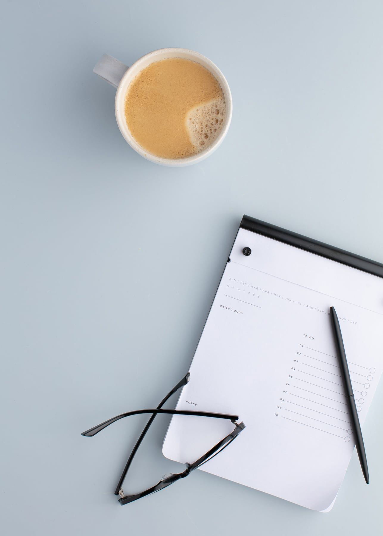 Coffee, notebook, pen, and glasses on a light blue desk in a minimalist workspace.