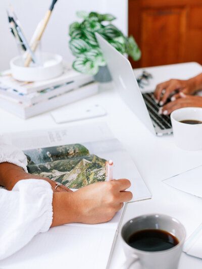 Person sketching on paper at a bright desk with coffee cup, books, and a laptop nearby