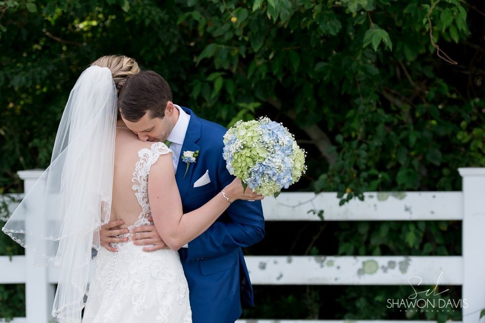 Bride and groom embracing beside a white fence, with a bouquet and lush green backdrop.
