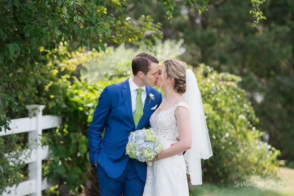 Bride and groom kissing in a garden, bride holding a white bouquet, groom in a blue suit
