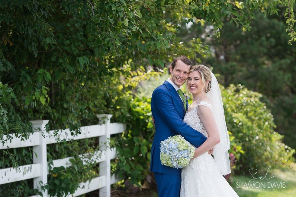 Newlyweds pose smiling by a white fence in a lush garden