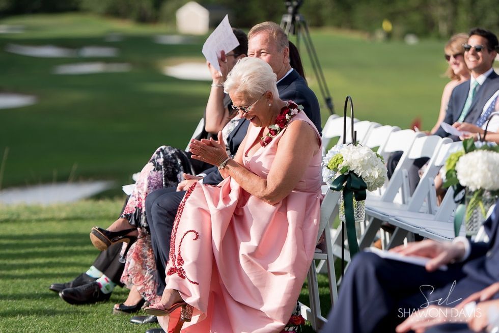 People seated outdoors at a formal event, one woman in a pink dress reading a program.