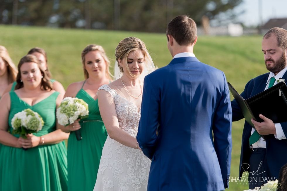 Bride and groom exchange vows outdoors, with bridesmaids in green and an officiant beside them.
