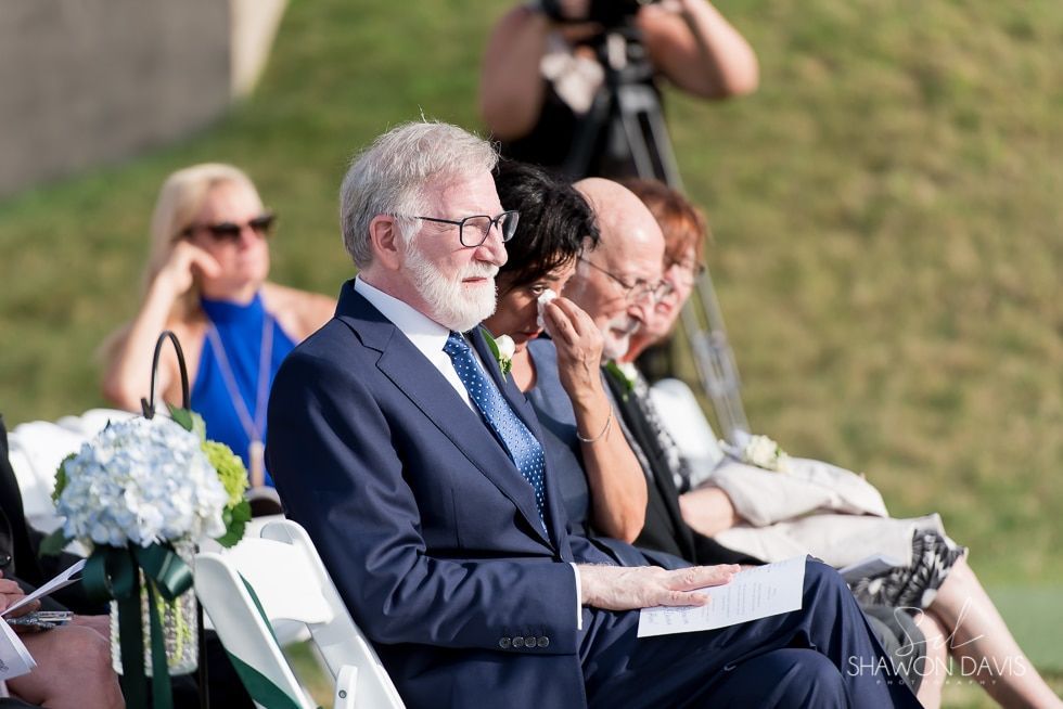 Guests seated outdoors at a formal event, including a man in a navy suit holding papers.