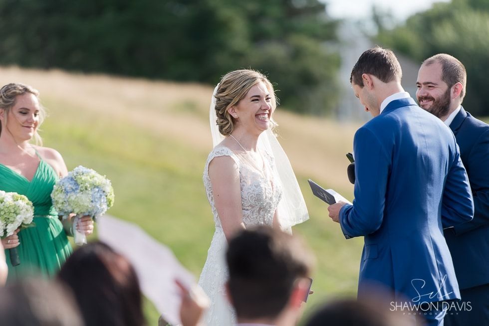 Bride smiling at outdoor wedding ceremony while groom exchanges vows with officiant nearby