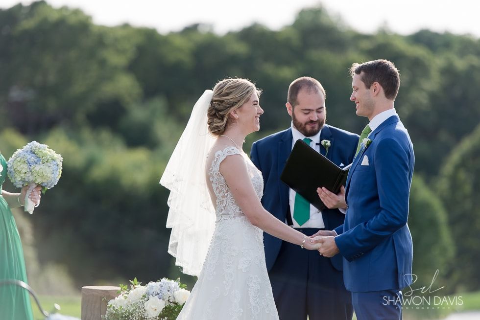 Bride and groom hold hands during an outdoor wedding ceremony officiated by a man in a suit.