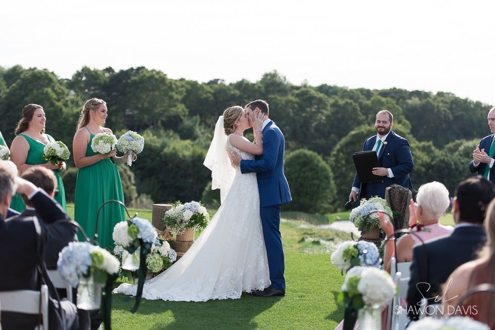 Bride and groom kiss at outdoor wedding ceremony on a lawn, surrounded by guests and white floral decor