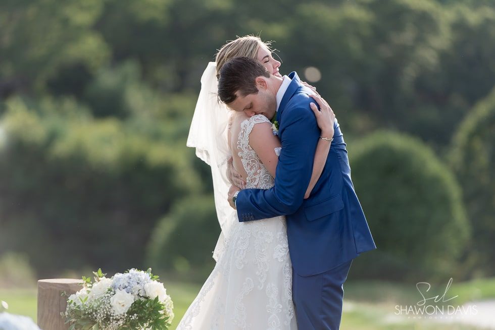 Bride and groom embrace outdoors in a sunlit garden, the groom in a blue suit and the bride in a white dress and veil