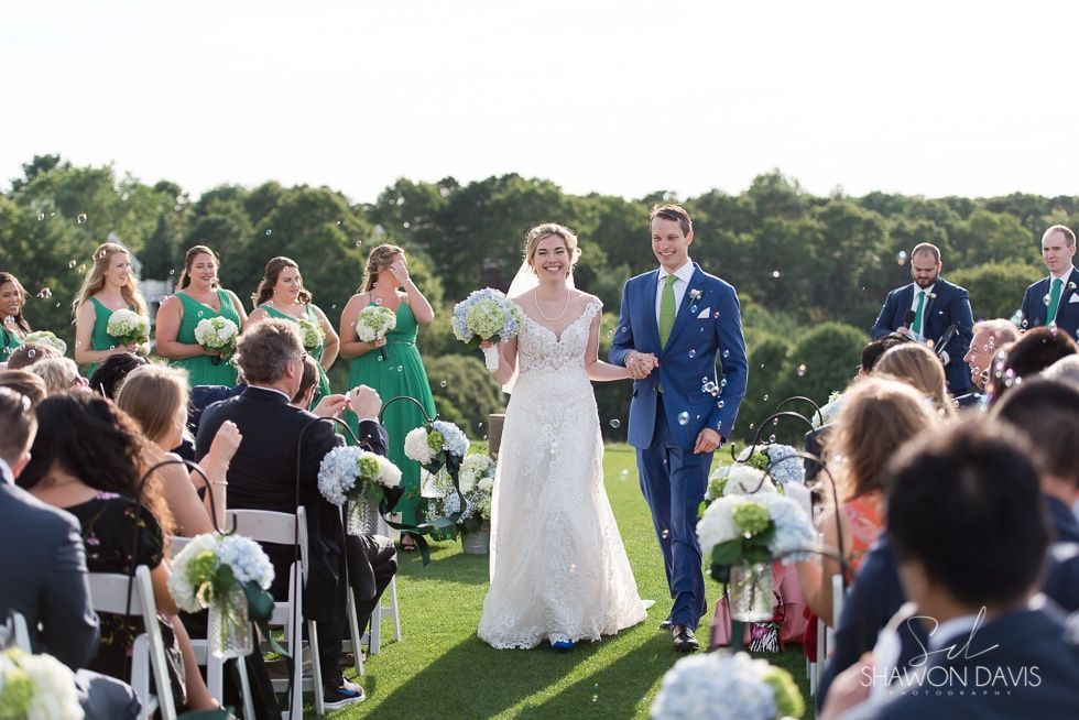 Bride and groom walking down an outdoor aisle at a wedding ceremony, guests seated on both sides