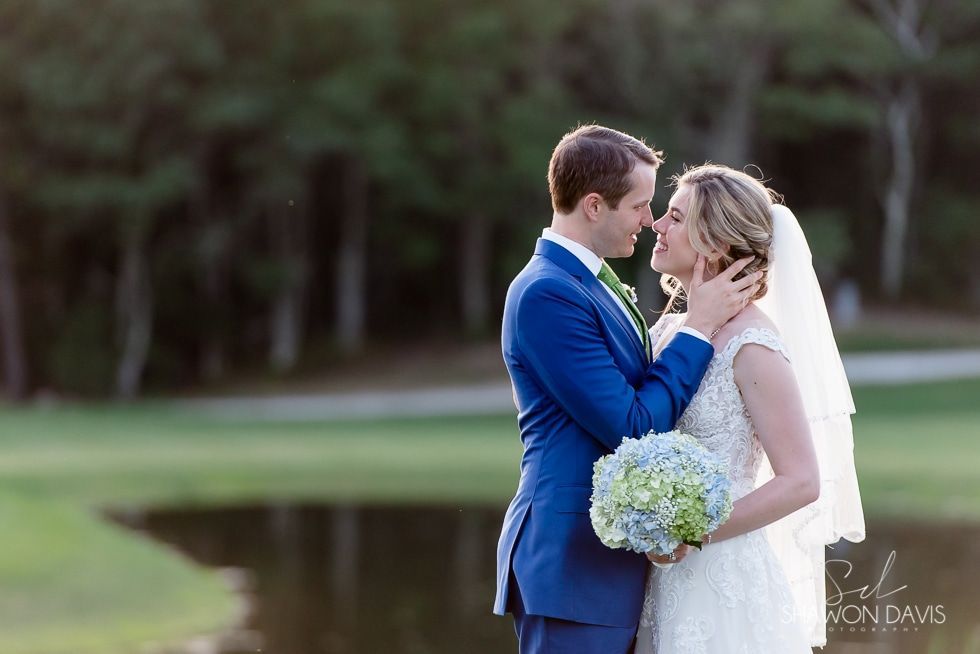 Bride and groom in blue suit and white dress share a kiss by a lake