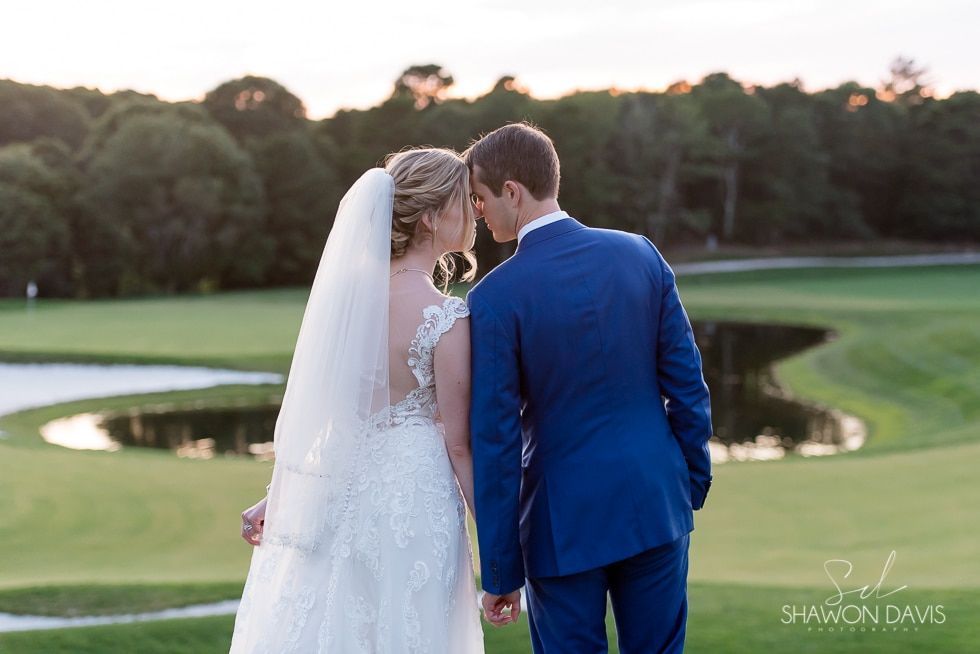Bride and groom in blue suit and white dress stand by a pond on a golf course at sunset